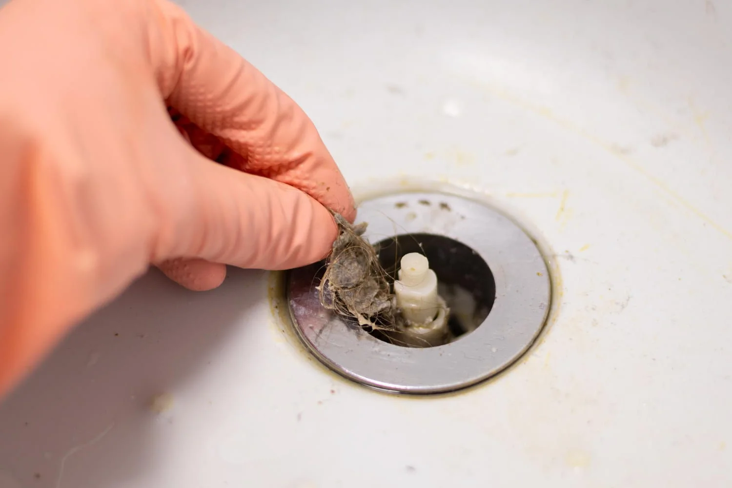 A gloved hand removes a clump of hair and debris from a bathroom sink drain.