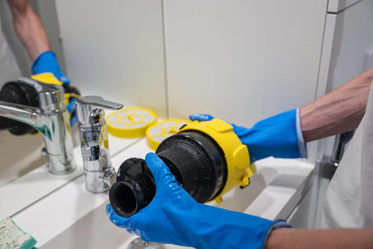 A person wearing blue gloves cleans a yellow and black vacuum filter under a bathroom sink.